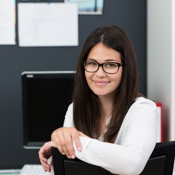 Friendly attractive young businesswoman wearing glasses leaning over the back of her chair at the office to smile at the camera